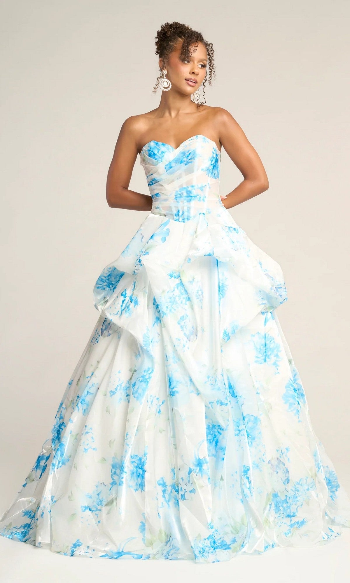 Woman wearing the Portia and Scarlett PS26083 long formal dress in white/blue, featuring a strapless corset bodice, blue floral pattern on white fabric, and a voluminous layered skirt, evoking an elegant floral organza look.
